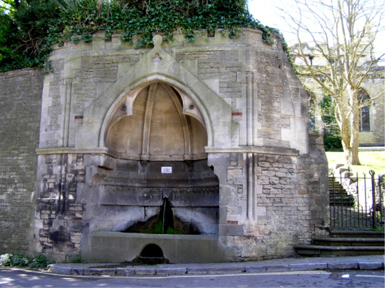 This well sits beneath a Gothic canopy beside the processional steps up to St John the Baptists Church.  The gates of the steps, which can be seen in the right of the picture, were locked when we visited.  It’s worth climbing up the steps to the right of the processional steps to look at the sculptured stations of the cross.

The well water flows from the mouth of a stone lions head into a bas