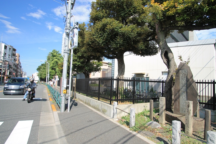 The monumental tablet under the tree on the right. The faculty of engineering of Tōkyō University beyond the fence.