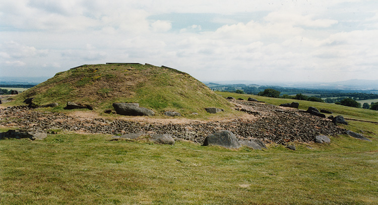 A general view of the outside of the site. The mound (concrete dome) represents the first stage of the cairn, and is thought to be the same diameter. Later the cairn was enlarged and this is represented by the flat area of stones, and it also is flanked by a circle of boulders.